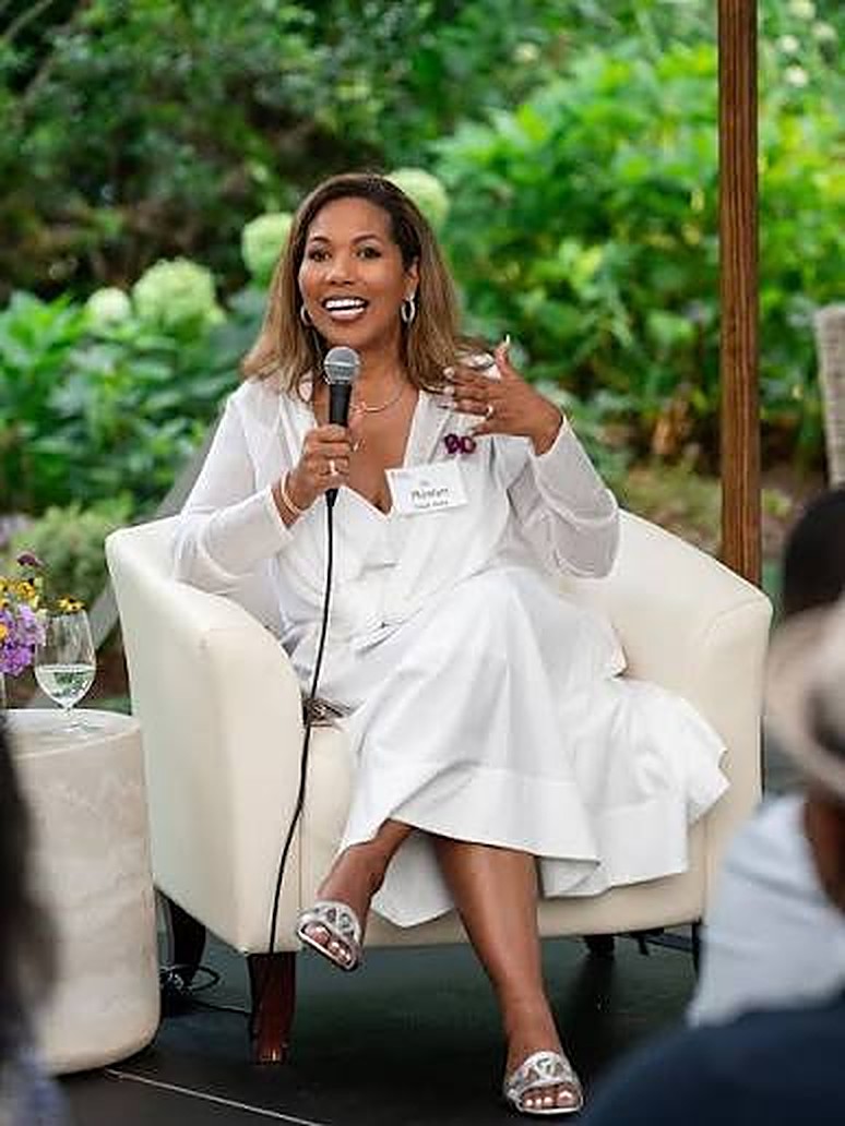 Dr. Roslyn Clark Artis seated in an armchair, speaking with a microphone, wearing a white dress, in front of a garden.